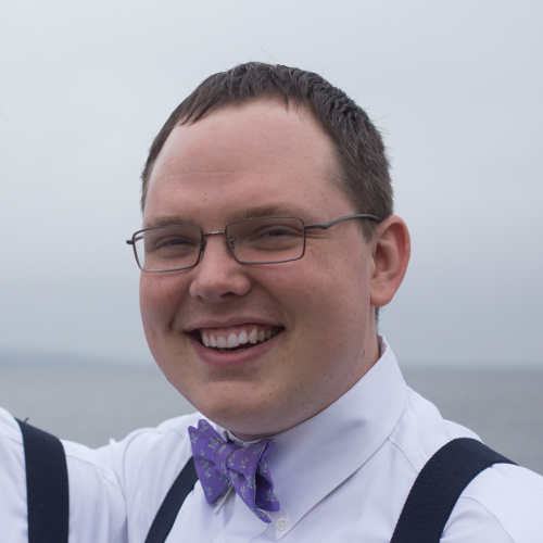 Photograph of Dan's head. Dan is a smiling white male in his 30s with short brown hair wearing glasses and a purple bow tie